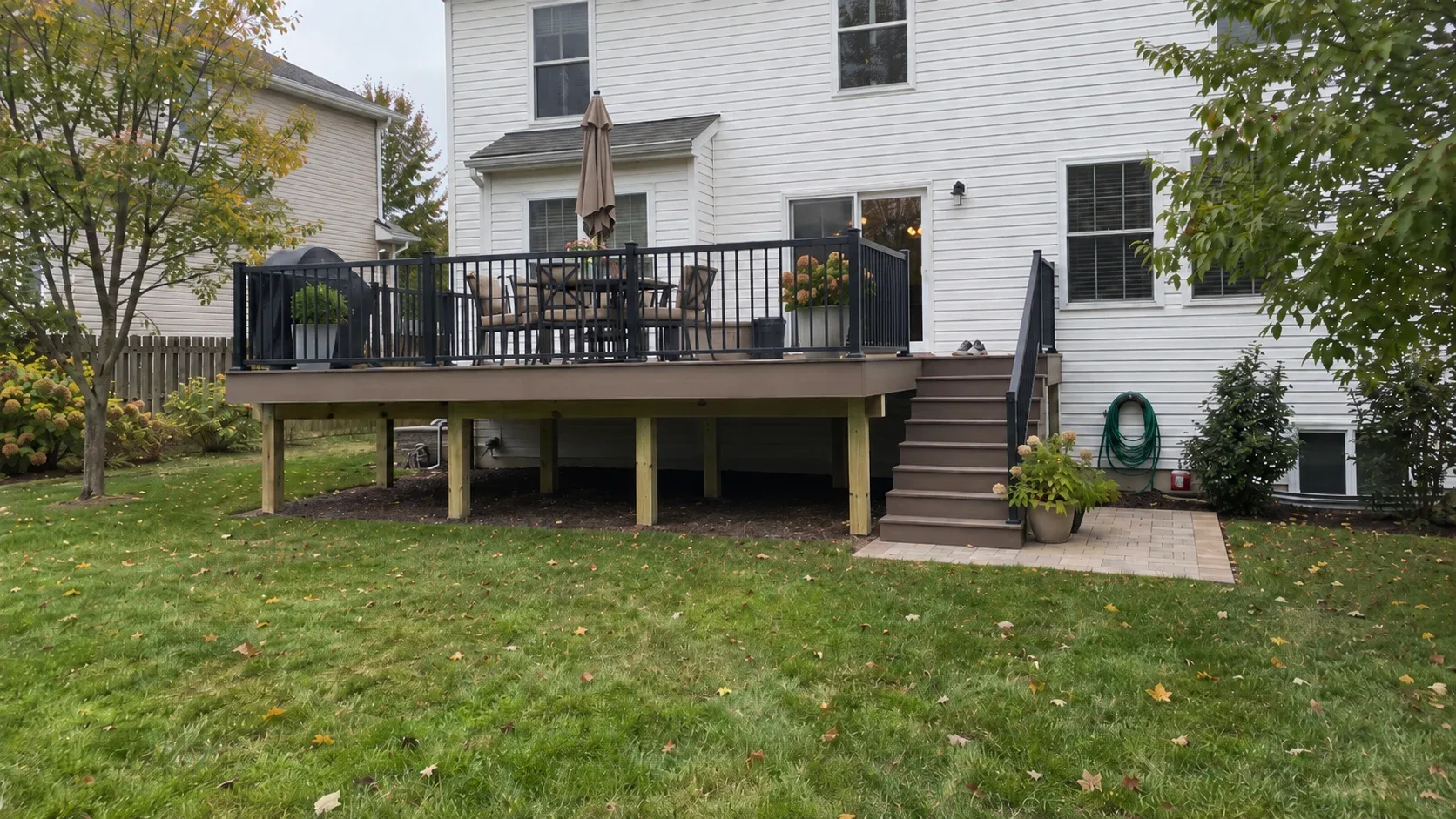 A composite deck lifted above a sloping lawn, black metal railings, steps running down to a paver landing and the frame visible in the open space underneath