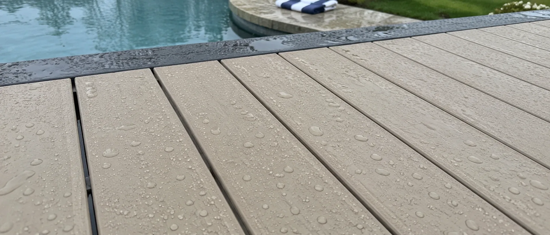 Water beading on pale capped boards at the edge of a swimming pool, with droplets standing proud and a blue pool beyond