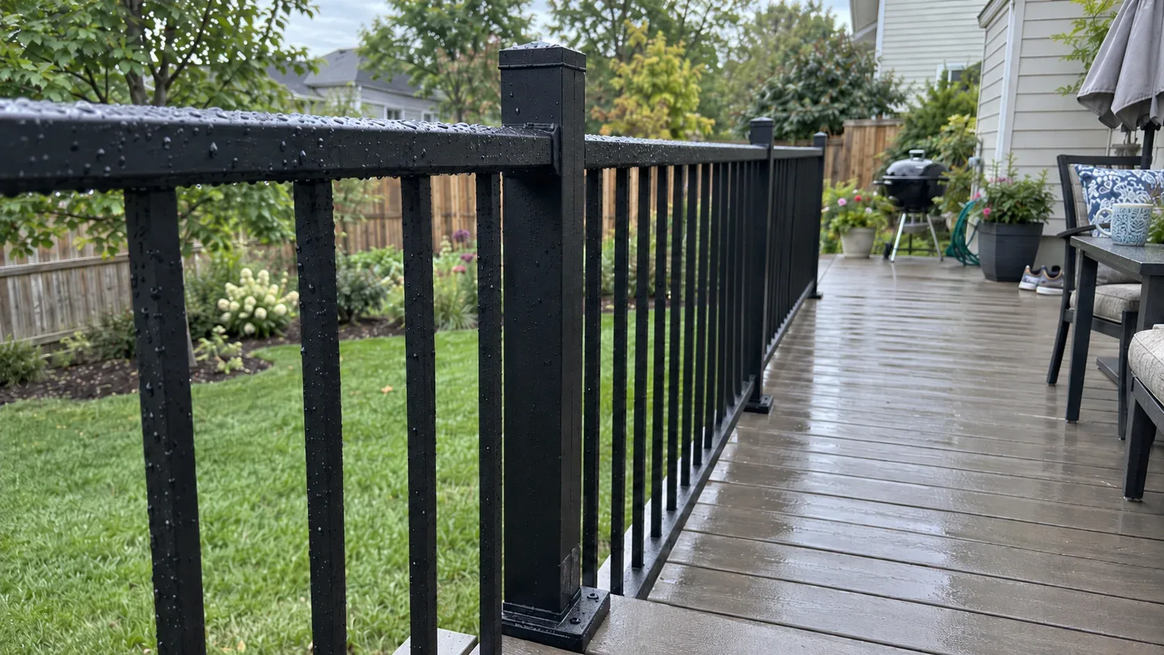 A dark composite deck edged with black aluminium railings and square balusters, a lawn and mature hardwood canopy behind